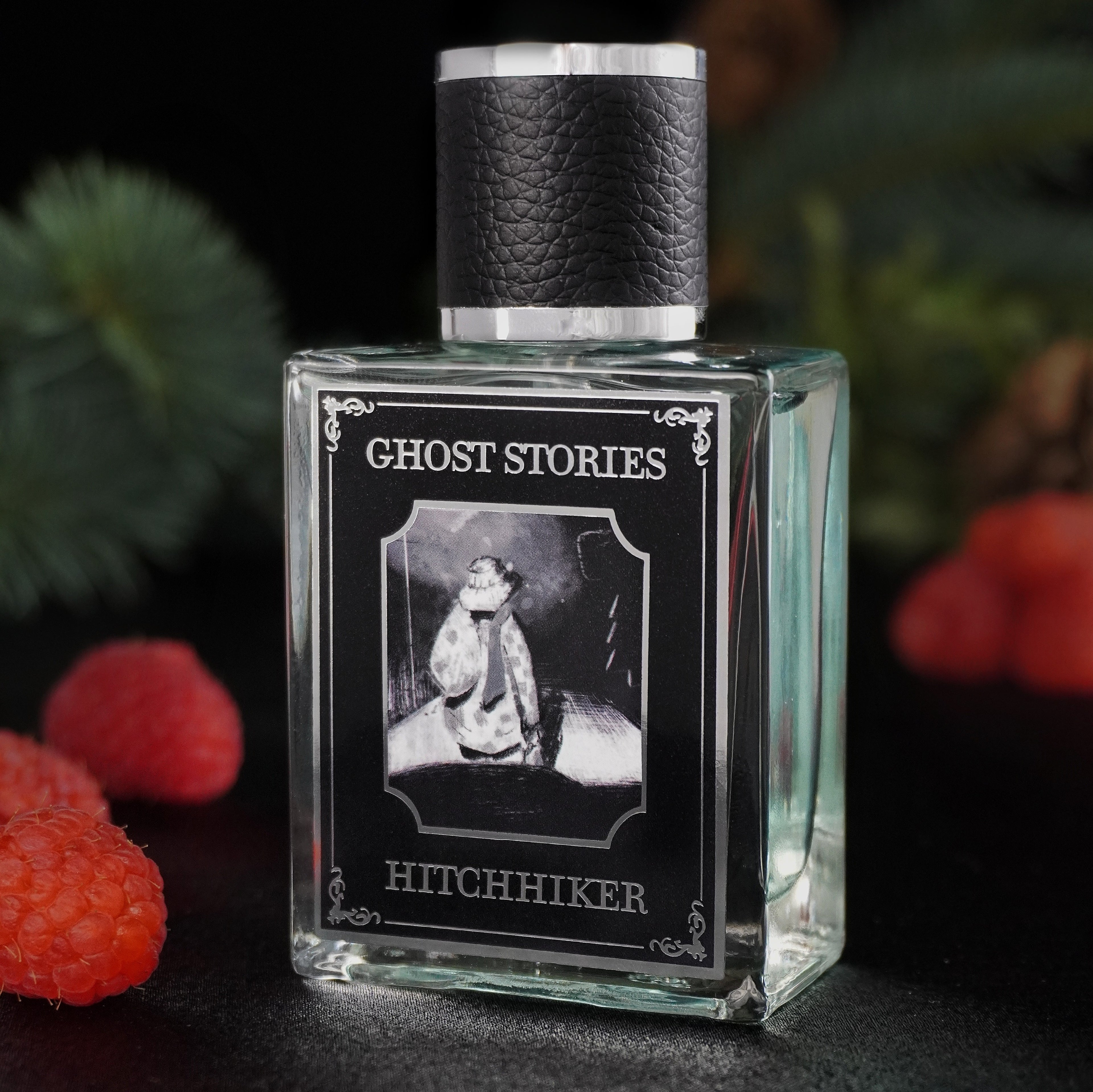 Perfume bottle labeled 'Ghost Stories Hitchhiker' with black cap, surrounded by raspberries and pine on a dark background.